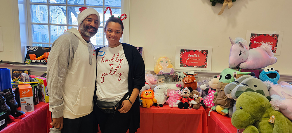 Two people smiling in front of a table full of toys for the holiday toy giveaway event